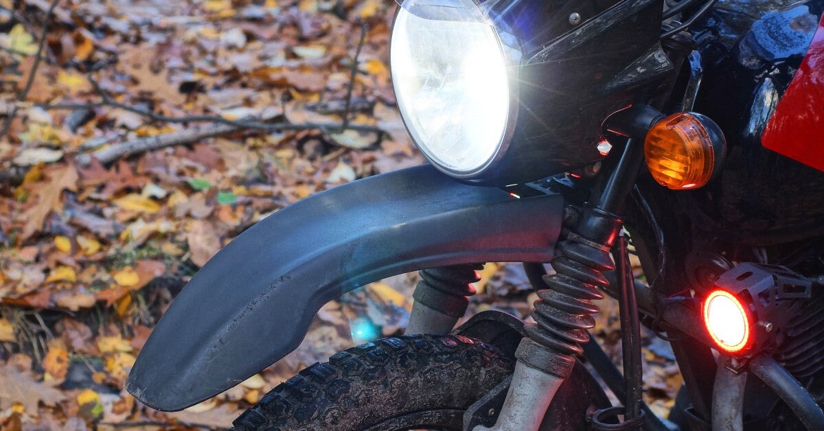 A motorcycle sitting in a woodland area surrounded by dead leaves. Its singular headlight casts a bright, white light ahead.