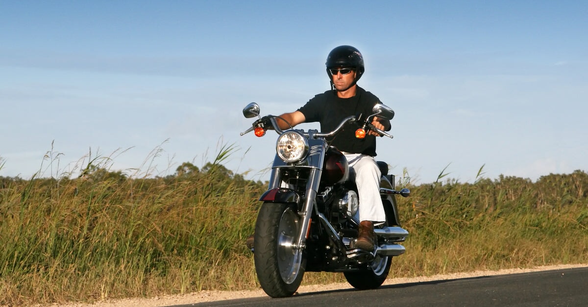 A man riding a motorcycle down the road, wearing a helmet and sunglasses. Fields of tallgrass border either side of him.