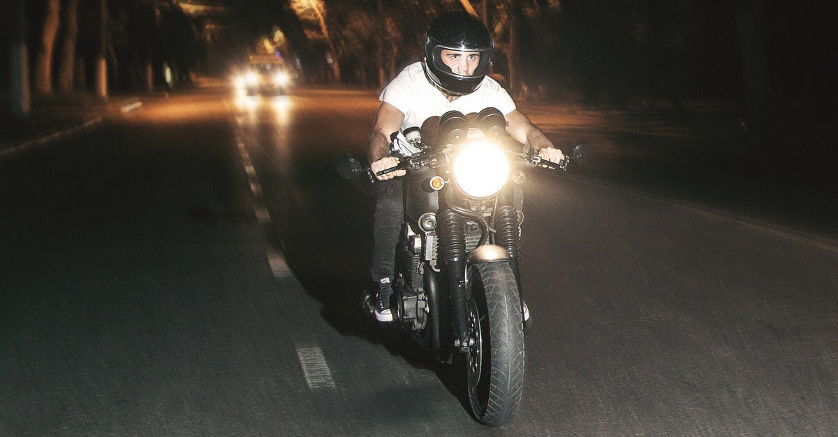 A man wearing a white tee shirt and a black helmet rides a motorcycle with a bright headlight down a dark road.
