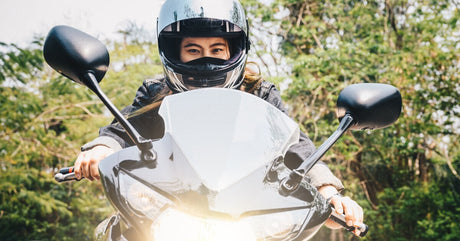 A close-up showing a young woman wearing a helmet riding a motorcycle that has a bright headlight on the front.