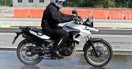 Somebody wearing an all-black outfit and a silver helmet rides a silver motorcycle down the road on a rainy day.
