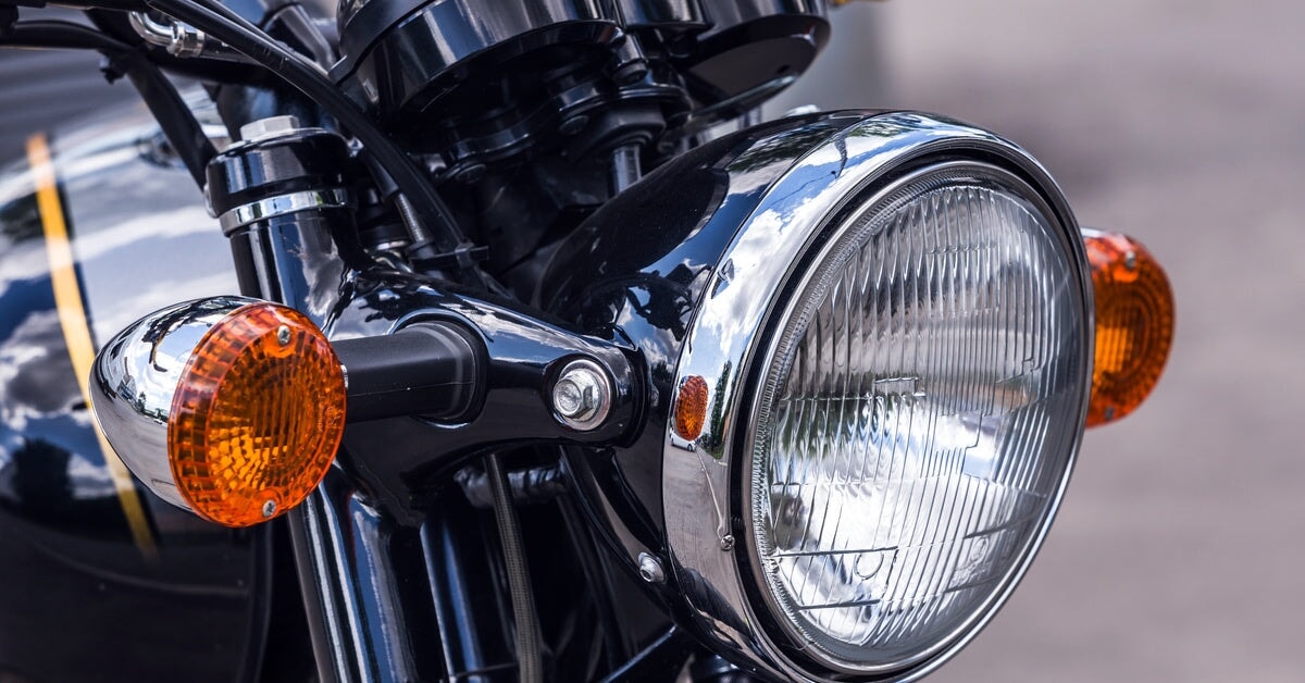 An extreme close-up showing a round, white motorcycle headlight that is attached to a dark colored motorcycle.
