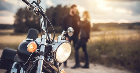 A close-up showing a motorcycle with an illuminated halogen headlight in front of a man and woman standing on the road.
