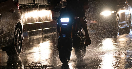 A motorcycle with a bright white headlight rides next to cars down the street on a dark, rainy night.