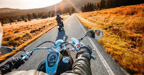 A close-up of a person wearing a leather jacket riding a shiny motorcycle down a country road behind another rider.