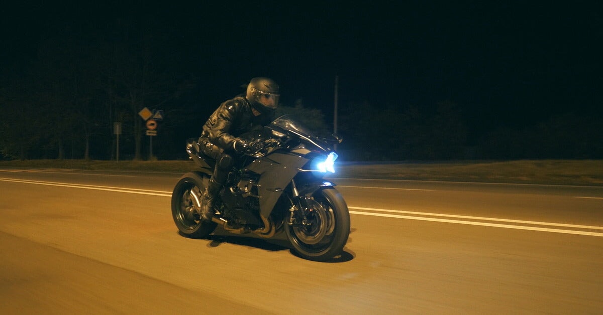 A man wearing an all-black, leather outfit rides a black motorcycle with a bright headlight down a street at night.