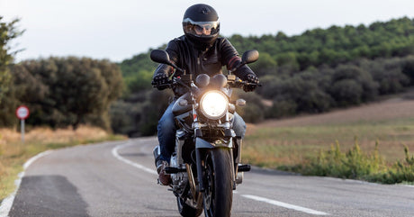 A man wearing a black helmet, leather jacket and gloves rides a motorcycle with a bright headlight down a country road.