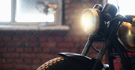 An older model of a motorcycle is parked with a brick wall and window in the background while it's headlight shines bright.