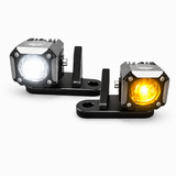 Two square off-road LED light units on black mounting brackets, one showing a white light and the other showing an amber/yellow light, on a white background.