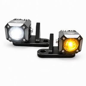 Two square off-road LED light units on black mounting brackets, one showing a white light and the other showing an amber/yellow light, on a white background.