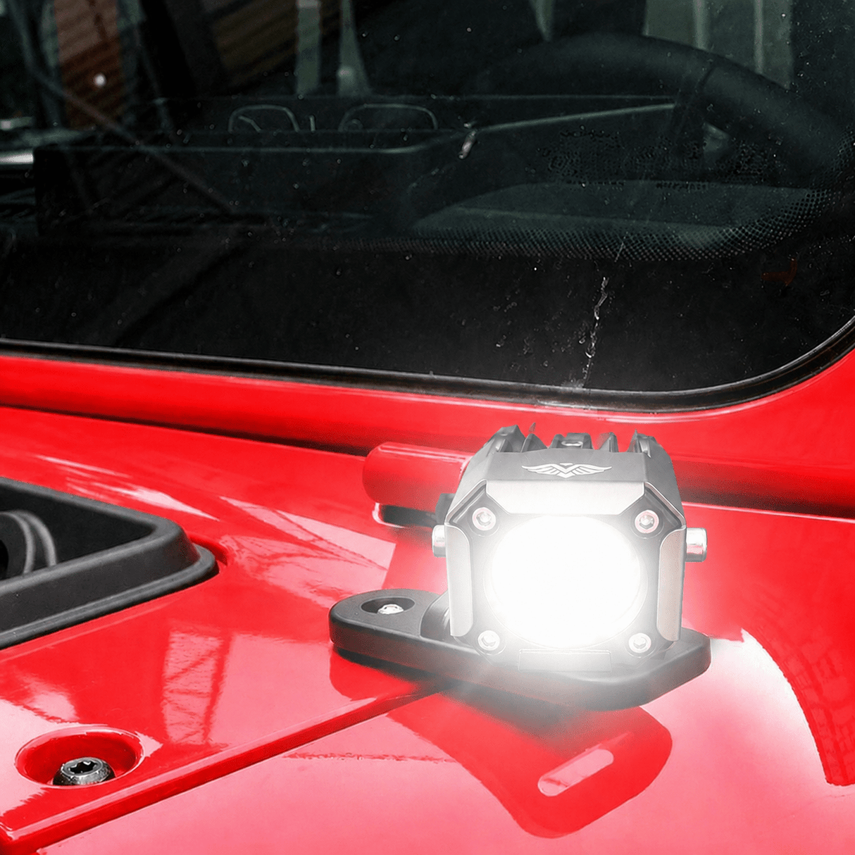 A small rectangular automotive light mounted on the glossy red hood of a vehicle, shown with its front illuminated. The light has a black housing and base with visible mounting screws.