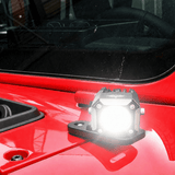 A small rectangular automotive light mounted on the glossy red hood of a vehicle, shown with its front illuminated. The light has a black housing and base with visible mounting screws.